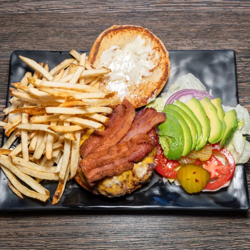 A bacon cheeseburger with avocado, tomato, onion, pickles, and fries on a black plate.